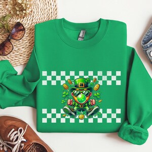 May include: Green sweatshirt with a checkered pattern and a cartoon leprechaun wearing a green hat, sunglasses, and holding a gold coin. The leprechaun is surrounded by shamrocks and gold coins.