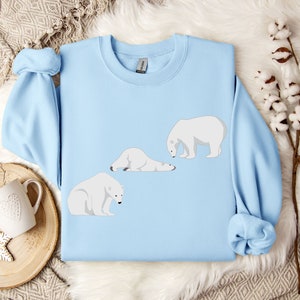 Peut inclure: Un sweat-shirt bleu clair avec un motif de trois ours polaires. Un ours est debout, un autre est couch&eacute; et le dernier est assis. Les ours sont tous blancs.