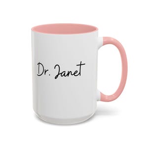 May include: White ceramic mug with a pink rim and handle. The mug has the text "Dr. Janet" written in black cursive.