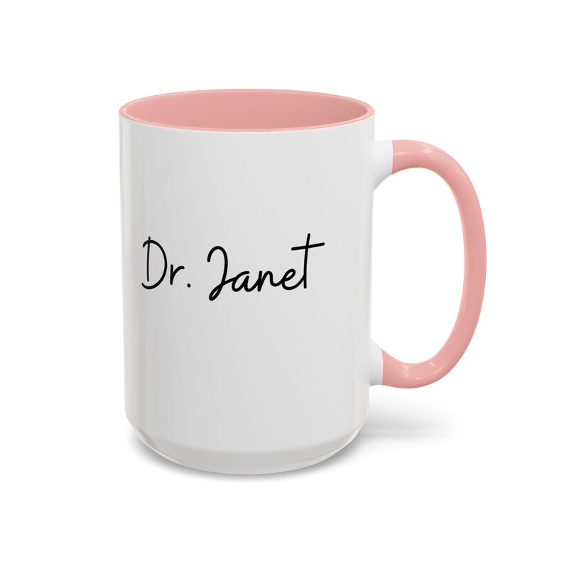 May include: White ceramic mug with a pink rim and handle. The mug has the text "Dr. Janet" written in black cursive.