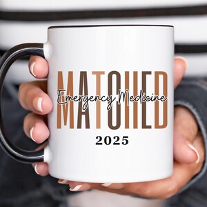 May include: White coffee mug with a black handle and black rim. The mug has the text "MATCHED Emergency Medicine 2025" printed on it in black and brown.