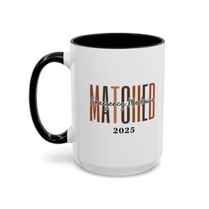 May include: White ceramic mug with a black rim and handle. The mug has the text "MATCHED Emergency Medicine 2025" printed on it in brown and black.