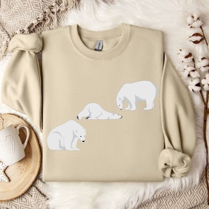 Peut inclure: Un sweat-shirt beige avec un motif de trois ours polaires blancs dans diff&eacute;rentes poses. Les ours sont tous tourn&eacute;s dans la m&ecirc;me direction, et le sweat-shirt a un col rond.