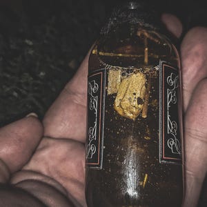May include: A dark amber glass bottle with a black label and ornate white and red accents. The bottle contains a liquid with a floating, golden-colored object. The bottle is held in a hand.
