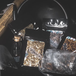 May include: An assortment of dried herbs and ingredients for spiritual practices. The image features small black pouches and a glass bottle with a cork, filled with various botanicals. The word "Wormwood" is visible on one pouch.