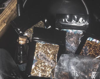 May include: An assortment of dried herbs and ingredients for spiritual practices. The image features small black pouches and a glass bottle with a cork, filled with various botanicals. The word "Wormwood" is visible on one pouch.