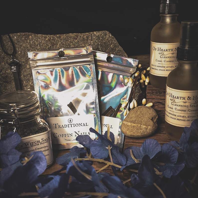 May include: An assortment of spiritual items, including bottles, jars, and pouches. The pouches are labeled "Traditional Coffin Nails." Bottles are labeled "Of Hearth & Cleansing." Blue flower petals and a small stone are also present.