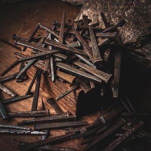 May include: A collection of weathered metal nails, showing signs of age and use. The nails, in various sizes and shapes, are scattered on a wooden surface, with some resting on a small wooden block. The color scheme is primarily brown and rust.