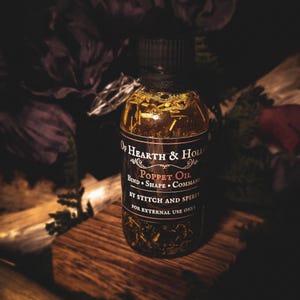May include: A clear glass bottle of Poppet Oil with a black cap, filled with herbs and oils. The label reads "Of Hearth & Hollow" and "Poppet Oil" with instructions. The bottle sits on a wooden block, with dark purple flowers in the background.