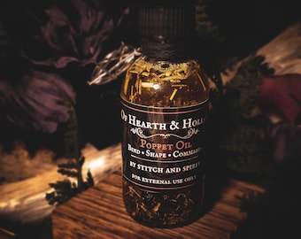 Peut inclure: Une bouteille en verre transparente d'huile Poppet avec un bouchon noir, remplie d'herbes et d'huiles. L'étiquette indique "Of Hearth & Hollow" et "Poppet Oil" avec des instructions. La bouteille est posée sur un bloc de bois, avec des fleurs violettes foncées en arrière-plan.
