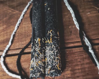 May include: Two black candles with a textured surface and gold and white accents. A white and black rope is draped across the candles, which are set on a wooden surface. The candles are cylindrical and appear handmade.