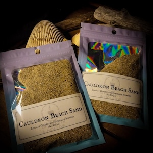 May include: Two pouches of Cauldron Beach Sand, each with a clear window revealing the sand. The pouches have a lavender top and a label with the text "Cauldron Beach Sand". A seashell and driftwood are in the background.
