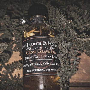 May include: A dark glass bottle with a black label featuring the text "Of Hearth & Hollow Cross Grave Oil." The bottle is wrapped with black twine and surrounded by green foliage. The label also includes the phrases "Decay, Ill Luck, Bind" and "For External Use Only."