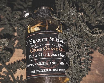 Peut inclure: Une bouteille en verre foncé avec une étiquette noire portant le texte "Of Hearth & Hollow Cross Grave Oil". La bouteille est enveloppée de ficelle noire et entourée de feuillage vert. L'étiquette comprend également les phrases "Decay, Ill Luck, Bind" et "For External Use Only."