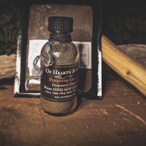 May include: A glass bottle filled with a brown powder, labeled "Of Hearth & Home Predator Grimoire." A silver snake charm wraps around the bottle. The label includes the text "Dominion, Fear, When Power Must Answer, Call the One Who Stands, For External Use."
