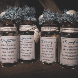 May include: Four glass jars with cork stoppers, twine, and decorative moss. Each jar has a label with text: Driftwood, Seaweed, Rosemary, and Moonstone. Seashells adorn the tops. The jars are arranged on a dark wooden surface.