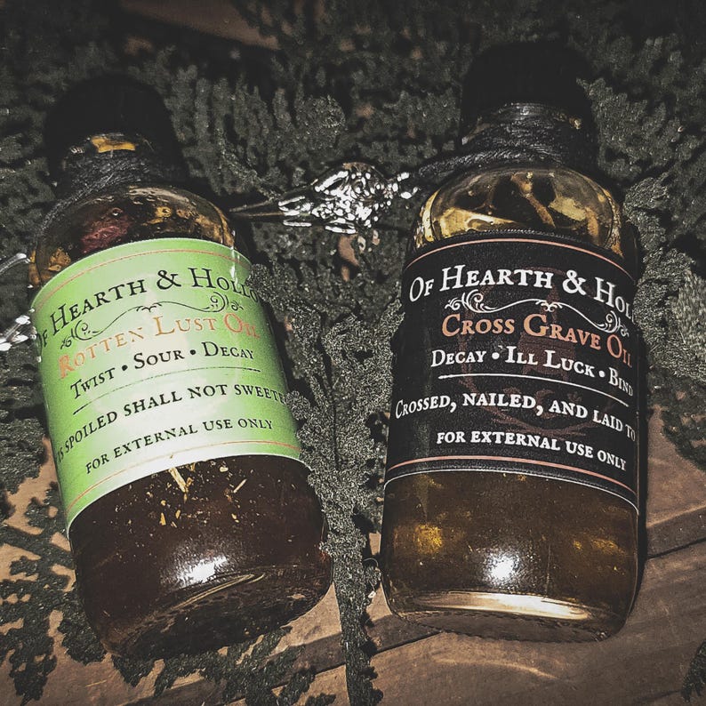 May include: Two glass bottles of Hearth & Hollow oils, one with a green label reading "Rotten Lust Oil" and the other with a black label reading "Cross Grave Oil." Both bottles are sealed with black caps and twine, with text that reads "For external use only."