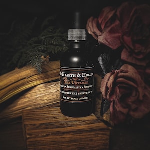 May include: A black bottle with a dropper, labeled "The Untamed" by Hearth & Hollow. The bottle sits on a wooden surface with dried flowers and foliage in the background. The text on the label includes "Sovereign" and "For external use only."