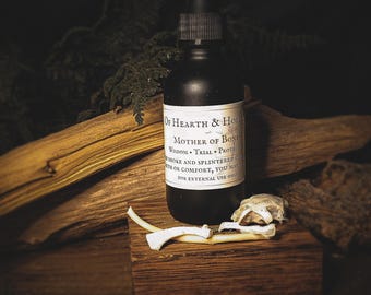 Peut inclure: Une bouteille noire avec un compte-gouttes, étiquetée "Mother of Bones", est posée sur un bloc de bois avec des fragments d'os. L'étiquette indique : "Of Hearth & Hollow, Wisdom, Trial, Protect, Smoke and splintered, for comfort, you may, for external use only."