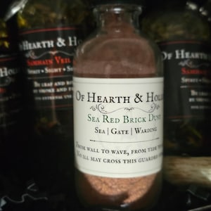 May include: A clear glass bottle filled with reddish-brown powder, labeled "Of Hearth & Hollow Sea Red Brick Dust." The label also includes "Sea | Gate | Warding" and other text. Other bottles are visible in the background.