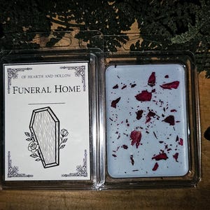 May include: A clear plastic clamshell package containing a Funeral Home scented wax melt. The wax is light blue with red rose petals. The label reads "OF HEARTH AND HOLLOW" and "FUNERAL HOME" with a coffin illustration.