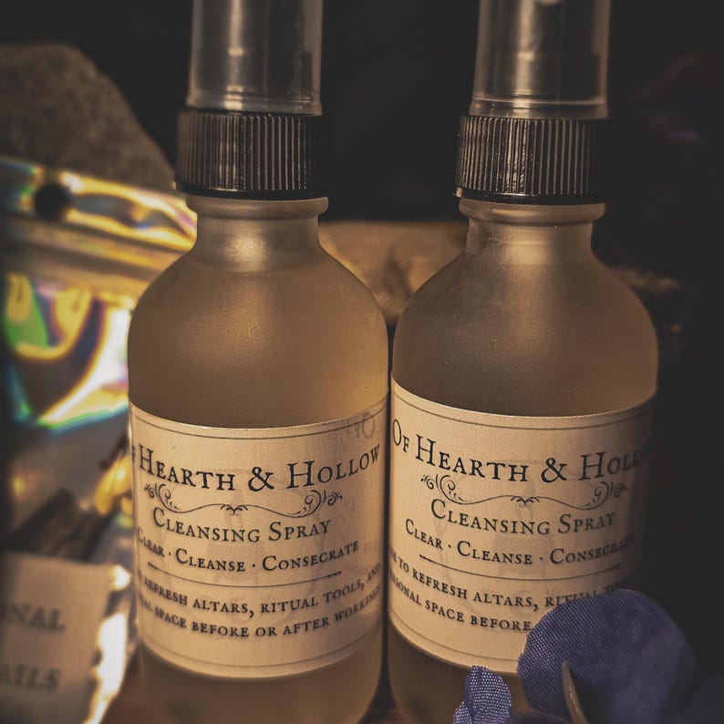 May include: Two glass spray bottles with dark metal spray tops. Each bottle has a label that reads "Hearth & Hollow Cleansing Spray". The bottles are filled with a light amber liquid. The labels also include the words "Clear, Cleanse, Consecrate".