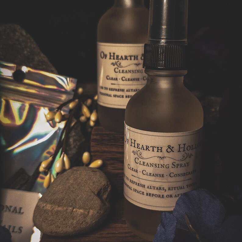 May include: Two brown glass bottles of "Cleansing Spray" with black spray nozzles. The labels read "Of Hearth & Hollow" and "Clear, Cleanse, Consecrate". A small rock and a bundle of yellow berries are in the foreground.