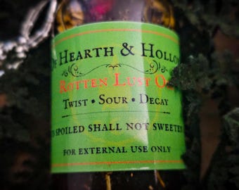 Peut inclure: Une bouteille en verre foncé avec une étiquette verte portant l'inscription "Hearth & Hollow" et "Rotten Lust Oil". L'étiquette comprend également les mots "Twist, Sour, Decay" et "For external use only."