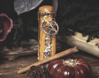 May include: A cylindrical orange candle with visible wax drips, embedded with dried herbs and botanicals, featuring a silver-toned pentagram charm attached near the top. A cinnamon stick, a small pinecone, and a dark red pumpkin sit nearby.