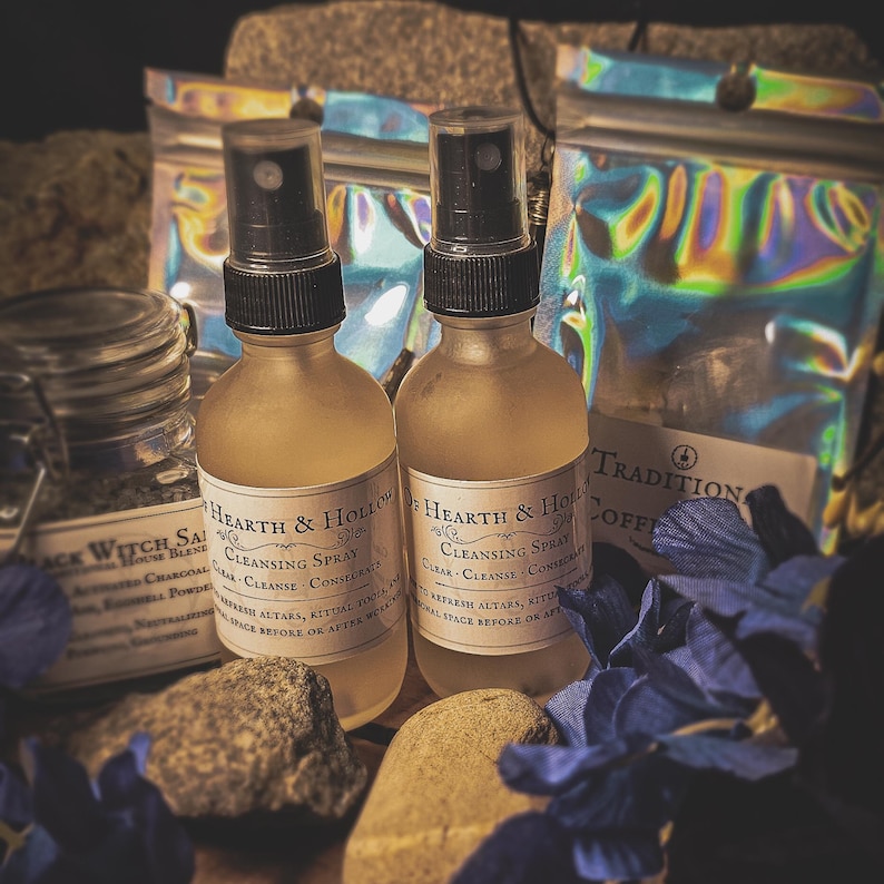 May include: Two glass spray bottles labeled "Hearth & Hollow Cleansing Spray" with black spray tops. The bottles are amber-colored, and the labels are cream-colored. Other products include a jar of "Black Witch Salt" and bags of "Traditional Coffee". The items are arranged with rocks and blue flowers.