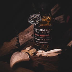 Peut inclure: Image sombre d'une bouteille d'"Huile de n&eacute;cromancie" avec un charme pentagramme. La bouteille, &eacute;tiquet&eacute;e "Hearth & Hollow", contient des herbes. Des fragments d'os et une grosse pierre sont dispos&eacute;s sur une surface en bois.