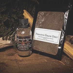 May include: A glass bottle and a black pouch containing "Predator Grave Dirt". The bottle, labeled "Hearth & Hollow", is filled with a granular substance. The pouch has a label that reads "Predator Grave Dirt" and "Use with Caution".