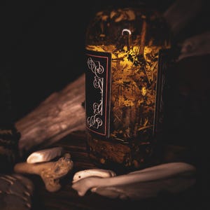 Peut inclure: Un bocal en verre transparent rempli d'herbes et d'un liquide dor&eacute;, avec une &eacute;tiquette noire orn&eacute;e d'une &eacute;criture blanche. Le bocal est entour&eacute; de bois flott&eacute; et de petites pierres claires, le tout sur un fond sombre.