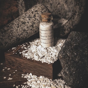 May include: A small glass bottle with a cork stopper, labeled "Hearth Shell Eggshell Powder," sits on a wooden block surrounded by eggshell fragments. The background includes a mortar and pestle and dark stones, creating a rustic aesthetic.