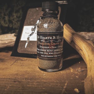 May include: A clear glass bottle filled with a granular substance, labeled "Predator Grave Dip" with text including "Dominion" and "Fear." The bottle is sealed with a black cap and tied with black string. A silver charm hangs from the bottle.