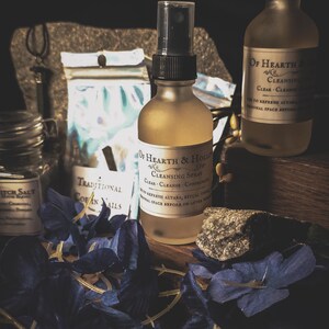 May include: A still life arrangement featuring two glass spray bottles labeled "Of Hearth & Hollow Cleansing Spray." Other items include a jar of "Witch Salt," a bag of "Traditional Covan Nails," and blue floral accents. The bottles have black spray tops.