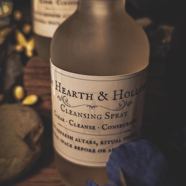 May include: A frosted glass bottle with a label that reads "Hearth & Hollow Cleansing Spray." The label includes the words "Clear, Cleanse, Consecrate" and "Refresh altars, ritual tools, space before or after." The bottle is on a wooden surface.