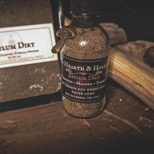 May include: A clear glass bottle filled with brown powder, labeled "Asylum Dirt" with a mushroom charm. The label reads "Of Hearth & Hollow" and includes text like "Obsession" and "Madness."