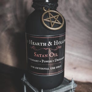 May include: A matte black glass bottle with a black twine-wrapped neck and a gold-toned pentagram charm. The label reads Hearth & Hollow, Satan Oil, Adversary Power Defiance, and For External Use Only in white and red text on a black background.