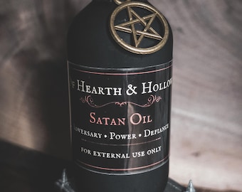 May include: A matte black glass bottle with a black twine-wrapped neck and a gold-toned pentagram charm. The label reads Hearth & Hollow, Satan Oil, Adversary Power Defiance, and For External Use Only in white and red text on a black background.
