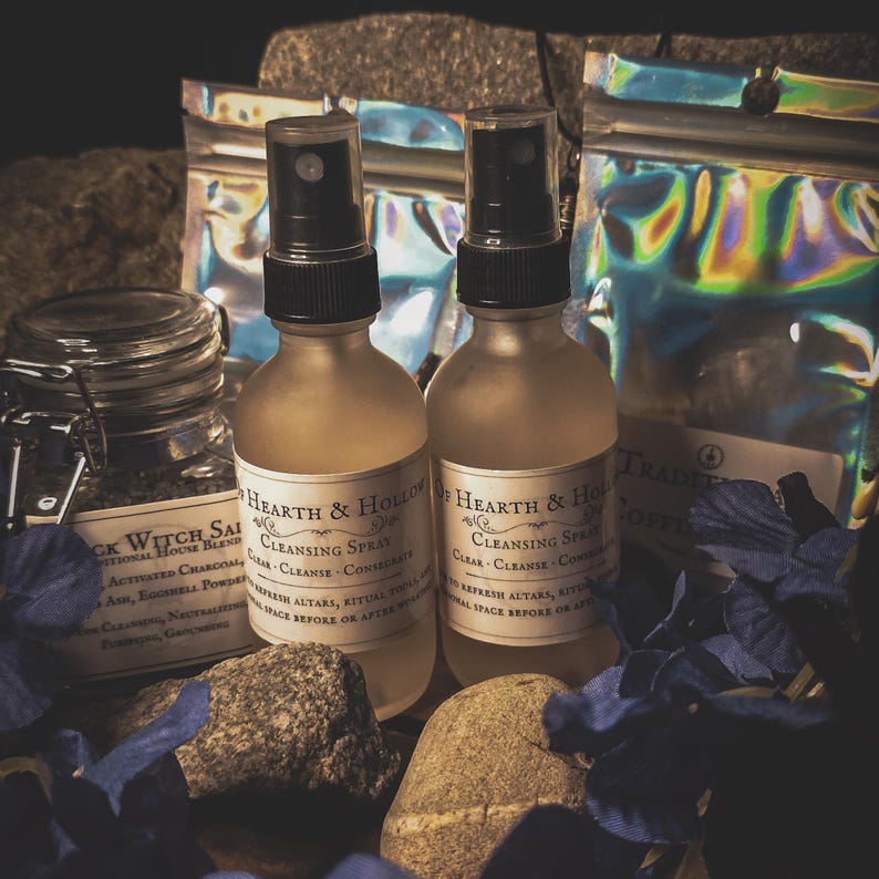 May include: Two frosted glass bottles of "Cleansing Spray" with black spray tops, labeled "Hearth & Hollow." A jar of "Black Witch Salt" and two iridescent pouches are also visible. The scene is decorated with rocks and blue floral accents.
