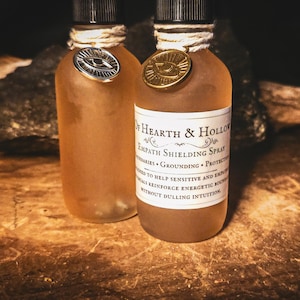 May include: Two amber-colored glass bottles with black caps and decorative twine. One bottle has a silver eye-shaped emblem, the other a gold one. The label reads "Of Hearth & Hollow Empath Shielding Spray".