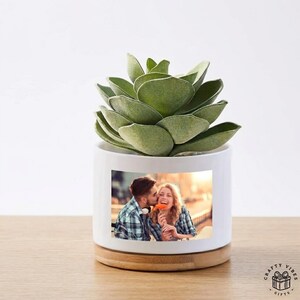 May include: A white cylindrical ceramic planter with a light wood base, containing a green succulent plant. The front of the planter features a rectangular color photograph of a couple sharing a moment, with the man kissing the woman's cheek.