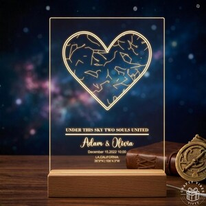 May include: A personalized LED night light featuring a heart-shaped constellation design. The light includes the text "UNDER THIS SKY TWO SOULS UNITED", names "Adam & Olivia", date, time, and coordinates. It sits on a wooden base.