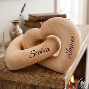 以下が含まれることがあります： 「Sophia」と「James」の名前が刻まれた木製のハート型の彫刻。 絡み合ったハートは明るい色の木でできており、木製の表面に置かれています。 背景には花瓶と本があります。