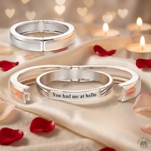 Puede incluir: Dos pulseras de metal plateado pulido con un acabado liso y redondeado. Una pulsera se muestra como un aro cerrado, mientras que la otra está parcialmente abierta, revelando el texto negro grabado "You had me at hello" en la superficie interior.
