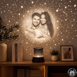 Può includere: Una lampada da proiezione cilindrica nera poggia su una superficie in legno, proiettando sulla parete un'immagine calda e luminosa di una coppia sorridente circondata da stelle scintillanti. La lampada presenta un design superiore forato.