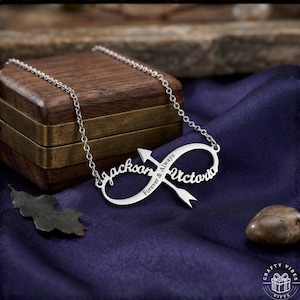 May include: A silver-colored necklace with an infinity symbol design. The necklace features the names "Jackson" and "Victoria" with the words "Forever & Always" and an arrow. The necklace is displayed on a blue cloth.