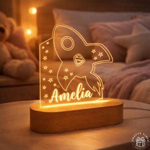 Puede incluir: Una luz de noche personalizada con un diseño de cohete y el nombre "Amelia" iluminado con un brillo cálido. Se asienta sobre una base de madera, creando un ambiente acogedor. El diseño incluye estrellas.