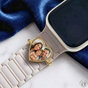 May include: A heart-shaped charm with a family photo, attached to a light grey smartwatch strap. The charm has a gold-coloured frame. The watch face is black. The background is a dark blue fabric.